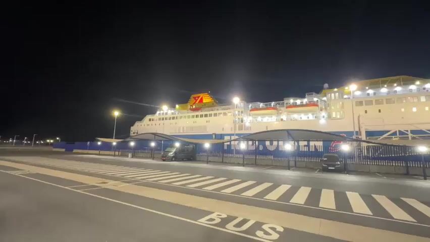 The video shows a large ferry docked at a terminal at night. The ferry is brightly lit with white and yellow lights. In the foreground, there is a striped pedestrian crossing with the word BUS painted on it. To the right of the crossing, there are covered parking spaces with lights, and a few vehicles are parked underneath. A blue fence separates the parking area from the ferry. The sky is dark, indicating it is nighttime.