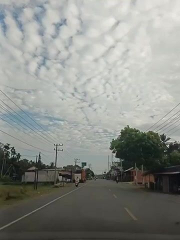 The video shows a view from a vehicle traveling on a paved road with a yellow line dividing the lanes. The road is lined with power poles and overhead wires. On the left side of the road, there are buildings and a truck. On the right side of the road, there are more buildings and trees. Several motorcycles are visible on the road. The sky is filled with scattered white clouds against a light blue background. The overall impression is of a rural or suburban road scene under a cloudy sky.