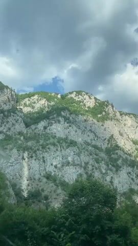 This is a vertical video of a rocky mountain slope covered in green vegetation under a cloudy sky. The camera is stationary and looking up at the mountain. The mountain is rugged and appears to be made of light-colored rock, with patches of green trees and bushes growing on its surface. The sky is mostly filled with dark, dramatic clouds, but there are also some patches of blue sky visible. The bottom of the frame shows a dense canopy of green trees.