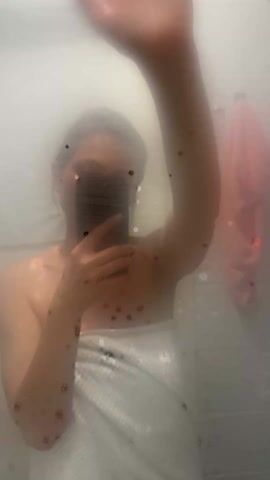 A person in a white towel is taking a selfie in a bathroom mirror that is steamed up. They wipe the steam off the mirror with their hand. A pink towel hangs on the wall behind the person. The person is holding a cell phone up to the mirror.