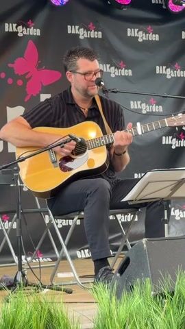 A man sits on a folding chair and plays an acoustic guitar and sings into a microphone. He wears a black short-sleeved shirt with subtle vertical stripes, black pants, glasses, and dark shoes. A dark strap goes from his guitar, over his shoulder and across his chest. He is on a stage in front of a black backdrop that has multiple instances of the word "BAKGARDEN" printed in white text, interspersed with pink butterfly graphics. There are pink stage lights visible at the top of the backdrop. In front of the man, there is a music stand holding sheet music. The foreground is filled with green blades of grass or similar foliage.