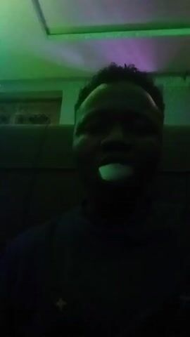 A low-angle, close-up shot shows a man exhaling a cloud of vapor, possibly from an e-cigarette or vape pen. The man has dark skin and short, dark curly hair. He is wearing a dark-colored shirt. The background is dark, with faint shapes of a sofa and what might be a window or light fixture visible. The scene is lit with shifting colors of green, blue, and purple, creating a moody atmosphere. The vapor obscures the man's face as it dissipates.