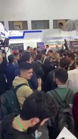 The video shows a crowded indoor scene, possibly at a convention or trade show. People are milling around, some in business attire, others more casually dressed. There are several cameras present, suggesting a media event or the presence of notable figures. Booths or displays with signage are visible in the background, including one labeled "evento". The overall impression is one of activity and potential interest or excitement.