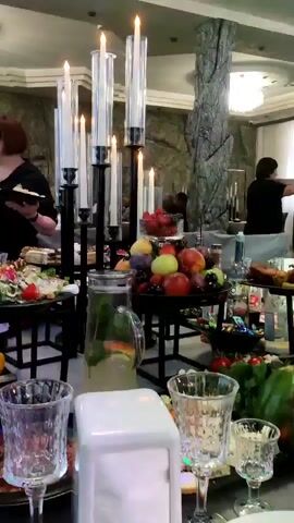 The video shows a formal gathering or party taking place indoors. A long table is laden with food and drinks, including fruit platters, salads, meats, and beverages. Tall candelabra with lit candles are placed on the table, contributing to the ambiance. In the background, several people are seen interacting, with some appearing to be dancing or mingling. The environment suggests a restaurant or event hall, with decorative elements like stone-like pillars and elegant ceiling lighting. The overall mood seems festive and social.