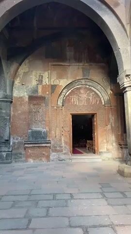 The video begins with a view of an arched entryway made of stone. To the left of the entryway is a stone carving embedded in the wall. The entryway itself is decorated with carved figures above the arch. Inside the entryway, an open doorway reveals a dimly lit interior with red carpet and wooden chairs. The camera then pans upwards to reveal a domed ceiling supported by columns, with an opening at the top allowing light to enter. The walls of the structure are made of stone with visible mortar and some areas of discoloration, suggesting age. The floor in front of the entryway is paved with large, square stones. The overall impression is of an ancient stone structure, possibly a church or religious building.