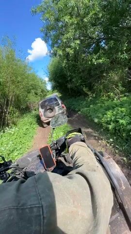 A first-person perspective shot shows an ATV driving on a dirt path through a lush green forest under a bright blue sky. In front of the ATV, a silver SUV is stuck in the mud. The driver of the ATV is wearing a green jacket and bright yellow gloves. A phone is mounted on the ATV handlebars. The scene is overgrown with dense foliage on either side of the narrow track. The SUV is covered in mud, indicating a recent struggle through the off-road terrain.