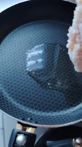 A close-up shot shows a piece of breaded chicken being placed into a frying pan filled with hot oil. The pan is black with a hexagonal pattern. The chicken is golden brown and appears to be coated in breadcrumbs. The oil is sizzling as the chicken makes contact. The scene is lit by overhead lighting, casting a slight glare on the pan.