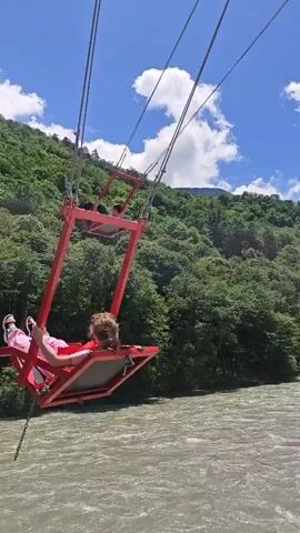 The video captures people enjoying large swings at an outdoor amusement site. The primary scene features individuals on red-framed swings soaring high above a wide, flowing river. In the background, densely forested green mountains rise under a bright blue sky dotted with fluffy white clouds. One person is prominently visible swinging towards the camera, while another is seen further out over the river.

A second perspective shows the swings from a different angle, revealing the substantial red and yellow support beams of the swing structure. Here, people are seated on the swings within what appears to be a park or attraction area, with parked cars visible behind a fence. Large green trees and distant mountains continue to define the picturesque landscape under the clear, sunny sky. The overall impression is one of an exhilarating outdoor experience in a mountainous, riverside setting, likely in Bzypta, Abkhazia, on a clear day. No visible text is present in the video.