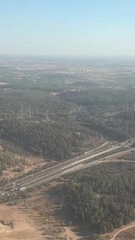 This aerial video shows a landscape featuring a highway interchange surrounded by a forest and agricultural fields. The highway, with multiple lanes and elevated sections, is the primary man-made structure visible, with cars traveling along it. The forest is dense and appears to be coniferous, with some scattered deciduous trees. The agricultural fields are a dry, golden color, suggesting a harvest season or dry conditions. In the background, a hazy horizon reveals more distant settlements and industrial structures, indicating a populated area beyond the immediate rural setting. Power lines and transmission towers are also visible, interspersed within the landscape.

The scene is captured on a clear day with bright sunlight, suggesting a mid-morning or early afternoon time. The sky is a pale blue with a slight haze. The overall impression is one of a developed, yet partially natural, landscape. The location is identified as Beit Nehemia, Israel, which provides context for the Mediterranean climate and typical vegetation seen in the video. No people are discernible in the video, and the focus is on the expanse of the land and infrastructure. The activity is simply vehicular traffic on the highway, indicating normal daily activity. There is no discernible text within the video itself.