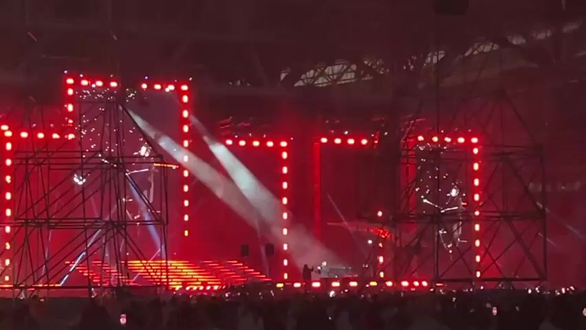 The video shows a wide shot of a concert or music festival stage illuminated by numerous red lights and several white spotlights. The stage features a complex structure of black metal scaffolding and platforms. At the center of the stage, there are several figures, likely performers or DJs, standing behind equipment. A large crowd of people is visible in the foreground, silhouetted against the stage lights. The overall atmosphere is energetic and vibrant, typical of a live performance event.
