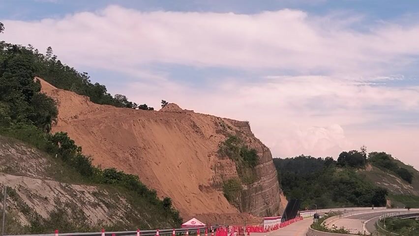 The video, filmed upside down, presents a moving view of a concrete highway, likely located in Kreet Paloh, Indonesia. The camera, appearing to be from a vehicle, advances along a gentle left-hand curve of the road.

On the right side of the main highway, a distinct line of red and white traffic cones borders a steep drop-off. Below these cones, a vast, exposed earthen slope descends into a valley. This slope displays visible geological layers and scattered green vegetation. Several red and white banners or flags are also discernible along this edge.

To the left, a concrete barrier separates the primary road from a lower-level road or ramp, which curves underneath. This secondary road also features clear lane markings. Beyond both roads, the landscape consists of green, tree-covered hills under a partly cloudy sky, indicating daytime.

The highway appears modern and well-maintained, characterized by clear yellow and white lane markings. No specific individuals are clearly visible, nor is any text on the distant banners legible. The scene highlights infrastructure development within a scenic, undulating terrain.