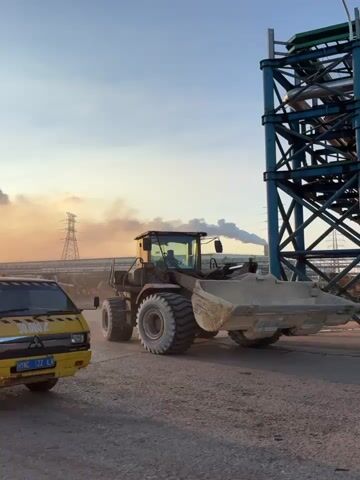 This media file captures a construction site in Keurea, Indonesia, during what appears to be sunset. The scene is dominated by heavy machinery, including a yellow pickup truck in the foreground with markings that read "HYNC" and "122". Behind it, a large loader with a prominent bucket and massive tires is partially visible, suggesting ongoing construction work. In the background, industrial structures, including a tall metal framework, power lines, and what might be storage tanks, are silhouetted against the sky.

The sky itself is a dramatic backdrop, showcasing a vibrant sunset with the sun a bright orb casting a warm, orange glow. Wisps of smoke or dust are visible, adding to the industrial atmosphere and possibly indicating ongoing activity at the site. The lighting suggests it is either early morning or late afternoon. The overall mood is one of industrious activity and the transition of day into night. The presence of heavy machinery and industrial infrastructure strongly indicates a construction or industrial area. The weather appears clear, with only a few scattered clouds softening the sunset's intensity.