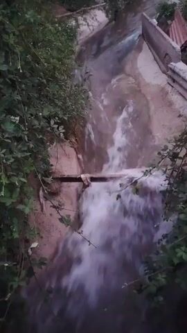 The video shows a stream of water flowing through a channel. The channel is lined with concrete and has some vegetation growing on its sides. There is a wooden plank that the water flows over, creating a small waterfall. In the background, there are trees and some structures. The water appears to be murky.