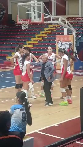 Here is a description of the video:

The video shows a group of young women in volleyball uniforms standing in a huddle on a basketball court. A man in a light-colored patterned blazer is speaking to them, gesturing with his hands. They appear to be in the midst of a timeout or coaching session. The court has red and blue lines, and there are basketball hoops visible in the background. The seating in the arena is red, yellow, and blue, and an advertisement for "Apuestas Royal" is prominently displayed. There are other individuals visible in the periphery, watching the scene unfold.