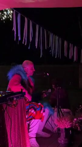 A bald person wearing a bright red hoodie with a blue furry collar and a patterned cloth draped over their lap is sitting in a chair. To their left is a DJ mixer on a table covered with a sparkly pink cloth. The person is looking down and to their right, towards another person who is holding a camera on a tripod. The background is dark, suggesting nighttime. Above them, there are strings of lights and colorful fabric streamers hanging from what appears to be a canopy. The scene is lit with pink and purple lights.