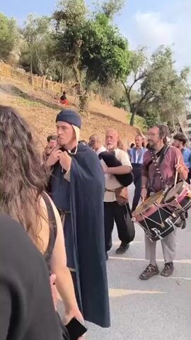 A group of people are walking down a dirt path lined with trees and a wooden fence. The path appears to be on a slope, with more people visible further up the hill. In the foreground, a person with long brown hair is facing away from the camera. The main focus of the video is a procession of musicians. One man in the center is playing bagpipes, another to his left is playing a stringed instrument, and a man to the right is playing a drum. They are all dressed in what appears to be medieval or Renaissance-style clothing. Several other people are following behind them. The sky is visible in the background, and the lighting suggests it is daytime, likely late afternoon.