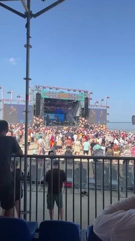 This video captures a live outdoor music festival from a standing perspective behind a black metal fence. The scene is dominated by a large stage set up on a sandy beach, with a clear blue sky overhead. A massive video screen displays a performance, showing musicians on stage. A large crowd of people, dressed in casual summer attire, is gathered in front of the stage, many holding umbrellas for shade. Colorful banners and stage lighting adorn the structure. The overall atmosphere appears to be energetic and festive, suggesting a sunny day at a beach concert.