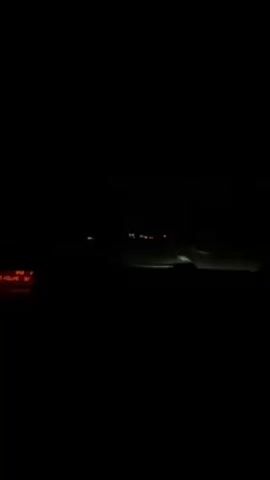 The video shows the dashboard of a car moving forward on an unlit road at night. The headlights of another vehicle are visible some distance ahead. The road has painted white lines marking lanes.
