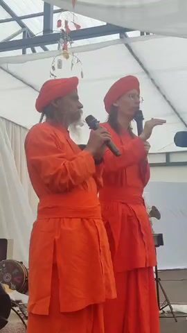 Two people in orange robes and head coverings stand under a white tent, each holding a microphone. The person on the left has a beard and is looking to their right, speaking into the microphone. The person on the right is wearing glasses and is looking forward, also speaking into a microphone. There are white curtains behind them, and some decorative items hang from the tent ceiling. A drum and a keyboard are visible to the left and right, respectively.