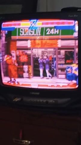 The video shows a person playing the video game Street Fighter on a Samsung CRT television. The game is displayed on the screen, showing two characters fighting in front of a building that appears to be a convenience store. The characters are engaged in combat, with one appearing to be in a crouched position while the other is standing. The television is sitting on top of a dark-colored wooden dresser. Cables are visible near the front of the TV. The camera angle is from slightly below the TV, looking upwards towards the screen.