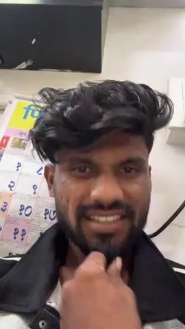 A close-up video shows a man with a beard and dark, thick, wavy hair looking directly at the camera. He makes a series of exaggerated facial expressions, initially pouting, then grimacing, smiling widely, and appearing intense or angry. Throughout the video, his hair is ruffled, possibly by a hair dryer, as a hand briefly enters the frame from the right to touch his hair. He also touches his own chin and neck at different points. In the background, a calendar is visible with "जून २०२३" (June 2023) written in Marathi, along with numbers for the days of the month. The setting appears to be indoors, possibly a salon or barber shop. The man's expressions convey a playful or reactive mood to the hair styling process.
