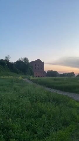 This is a vertical, low-angle, outdoor video taken at dusk. The scene depicts a rural landscape with a dirt path winding through a grassy field. In the background, a large, old brick building, appearing somewhat dilapidated, sits on a hill surrounded by trees. The sky is a gradient of pale blue at the top, transitioning to soft oranges and pinks near the horizon, suggesting the last light of sunset. The camera pans slowly to the right, following the path. There are no people or animals visible in the video.
