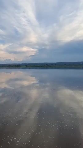 A vertical video displays a calm body of water reflecting a cloudy sky. The water is dark and still, with small white bubbles on its surface. The sky above is a mix of blue and white clouds, with some darker gray clouds present. A distant, dark horizon line separates the water from the sky, suggesting a landscape of rolling hills or distant trees. The reflection in the water is a mirror image of the sky, creating a symmetrical and serene scene. The lighting suggests it may be dawn or dusk. There are no people or objects visible in the frame.
