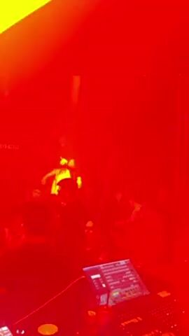 The video shows a live performance in a dark, red-lit venue, likely a club. On stage, three figures can be seen in the spotlight with microphones, apparently rappers. Below the stage, the backs of the audience members are visible, some are moving and interacting. In the foreground, a soundboard sits with its lit screen displaying numerous buttons and controls. The overall atmosphere seems energetic and club-like.