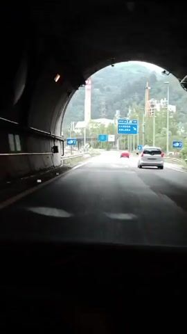 The video captures a view from inside a vehicle exiting a tunnel onto an open road. The road ahead is paved and has visible lane markings. There are several cars on the road, including a red car and a white car. On the right side of the road, there are signs indicating directions and distances. In the background, a tall, striped industrial chimney is visible, rising against a backdrop of green, misty mountains. The sky is overcast. The overall environment appears to be an industrial area near a mountainous region, likely on a cloudy day.