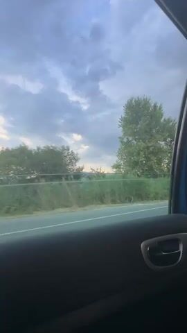 The video shows a view from inside a car, looking out the passenger window. The scene outside is a rural landscape with trees and vegetation. A road is visible running horizontally across the frame, with power lines and a power pylon suggesting the area is along a transport route. The sky is filled with dramatic, cloudy formations, with hints of sunlight breaking through in the distance, indicating it is either early morning or late afternoon. The overall mood is serene and contemplative, perhaps of a journey. There is no visible text. The location is identified as Kovachevo, Bulgaria.
