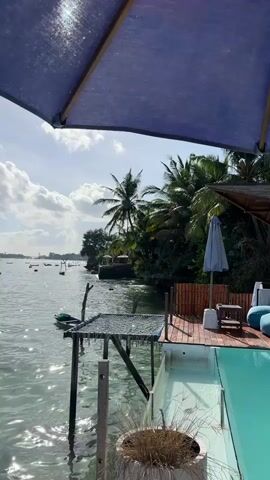 A wide shot captures a serene waterscape under a bright sky, framed by the deep blue canopy of a large umbrella. The water, shimmering with sunlight, stretches towards a distant shoreline dotted with lush palm trees and vegetation. Several small boats are scattered across the water, some appearing anchored. In the foreground, a wooden deck area is visible, featuring a clear blue swimming pool and a small table with seating. A bamboo fence runs along the edge of the deck, and a potted plant with dry grass sits beside a wooden post. A small, rickety structure with netting hangs over the water, supported by posts. A gentle breeze seems to be present, causing ripples on the water's surface and a slight sway in the palm fronds. The scene evokes a tranquil, tropical atmosphere.