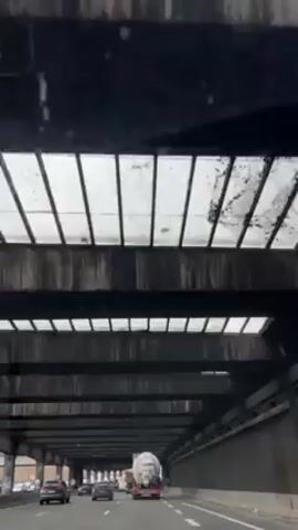 The video shows a view from inside a vehicle moving through a tunnel. The camera is pointed upwards towards the ceiling of the tunnel, which has a series of rectangular openings covered with translucent panels, allowing some light to filter through. The tunnel structure is made of dark, possibly concrete, material. Below the tunnel ceiling, traffic is visible, including cars and trucks moving in the same direction as the camera vehicle. The overall impression is of a functional, somewhat industrial environment.