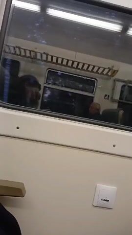 The video shows the interior of a train carriage. Through a window, two passengers are visible in reflection. One passenger, with dark hair, is leaning forward. Another passenger with lighter hair is seated further back. The train interior is light-colored with overhead luggage racks. A USB charging port is visible on the wall of the carriage. The reflection in the window shows trees and a blue sky, indicating movement.