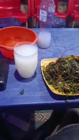 On a blue plastic table sits a plate of cooked greens, a glass of white liquid, a half-empty bottle of water, and a black electronic device. An orange plastic basin also rests on the table. The greens appear to be chopped and include some red pieces. The glass is about half-full. Red plastic chairs are visible behind the table. A person’s legs, clad in denim jeans, are seen beneath the table, resting on a patterned surface.