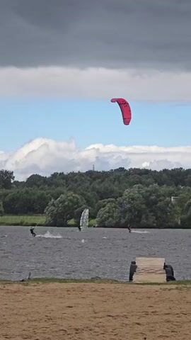 The video shows a wide shot of a body of water with several kiteboarders and one windsurfer. The sky is partly cloudy with dark clouds in the upper left corner and lighter clouds and blue sky in the upper right. A large red kite is prominently visible in the air. In the middle ground, there are trees lining the far bank of the water. In the foreground, there is a sandy beach with a wooden structure. There are at least three individuals visible on the water, engaged in their respective sports. One person on the left is kiteboarding and appears to be in motion, creating a spray. Another person is windsurfing with a sail visible. A third person is further to the right, also kiteboarding. On the beach, two figures are partially visible behind the wooden structure. The overall scene depicts an active watersports environment.