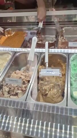 The camera pans across a display case filled with different flavors of ice cream. The display case is made of stainless steel and glass. The flavors include tiramisu, kinder bueno, pegauza, mint chocolate chip, stracciatella, forest berries, coconut, white chocolate and almanca, and passion fruit. A person reaches into the display case with an ice cream scoop and serves ice cream into a cone.