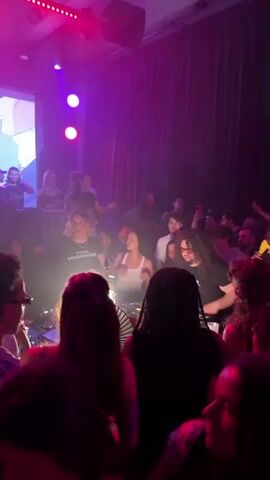 A crowded club scene is illuminated by pink and purple lights, with a projector screen displaying an animated face in the background. People are packed together, dancing and enjoying the music. Two DJs stand behind a table with electronic music equipment, operating the controls. One DJ is wearing a black t-shirt with white writing, while the other wears glasses. A woman in a white top dances between them. The crowd is diverse in age and appearance, with some holding phones up to record or take pictures. There is an energy and excitement in the room as people interact and move to the music.