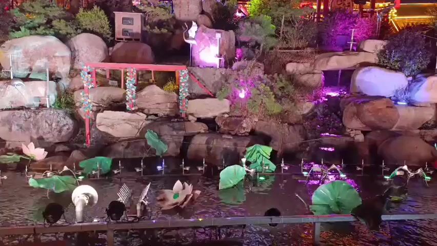 This video displays an outdoor nighttime scene with a decorative waterfall and pond area. The landscape is adorned with rocks, greenery, and artificial lotus flowers floating on the water's surface. Several artificial lights, some colored, illuminate the scene from various angles. In the background, there is a traditional East Asian architectural style building with glowing red lanterns. Above this, a large Ferris wheel is visible, also lit up with colorful lights. Two towers flank the Ferris wheel, each displaying text that appears to be in a Cyrillic alphabet. A solitary figure, seemingly dressed in light-colored clothing, stands on a raised platform near the top of the rock formation, possibly performing or posing. The overall ambiance is festive and theatrical, suggesting a park or entertainment venue.