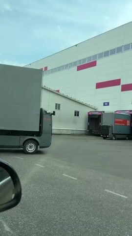 The video shows a gray EVOCARGO truck in front of a large warehouse. The truck is driving on an asphalt lot. The warehouse is white with pink stripes. The EVOCARGO truck is moving, and there are other trucks nearby. The warehouse has numbered loading docks. The video is shot from inside a car.