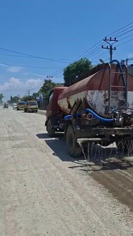 A red and white tanker truck is driving down a dusty dirt road, spraying water from a bar at the back of the truck onto the road surface. The truck has a large cylindrical tank on the back, with blue hoses connected to it. There are other vehicles on the road, including a yellow truck and some smaller cars. The road appears to be under construction or maintenance. The sky is blue and clear, with some power lines visible in the background. The overall impression is of a construction or road maintenance operation in a rural or semi-urban area.