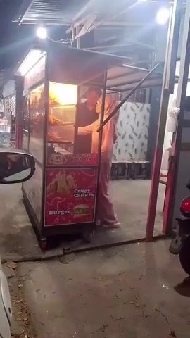 A person wearing a headscarf and light pink clothing stands behind a food cart. The food cart has a sign with "Crispy Chicken" and "Burger" visible. The cart appears to be cooking food, with flames and heat radiating from inside. Another person in a dark outfit walks past the cart. The scene is outdoors at night, with artificial lighting illuminating the food cart and surrounding area. The environment appears to be a street or walkway, with a car visible in the foreground.