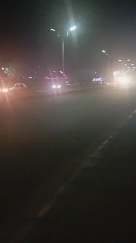 The video depicts a night scene on a road with cars and streetlights. The camera appears to be handheld and low to the ground. Cars pass by with bright headlights that illuminate the road. Streetlights are visible on either side of the road. The overall lighting is dim due to it being nighttime. There's no clear indication of people in the video. The surface of the road appears to be asphalt.