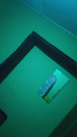 A video shows a room with green walls. In the center of the frame, a rectangular mirror is affixed to the wall. The mirror reflects a patterned object, possibly a piece of fabric or paper. The camera pans around the room, revealing a dark doorway and a window with blinds. The lighting in the room appears to be blue or green. There are no people visible in the video.