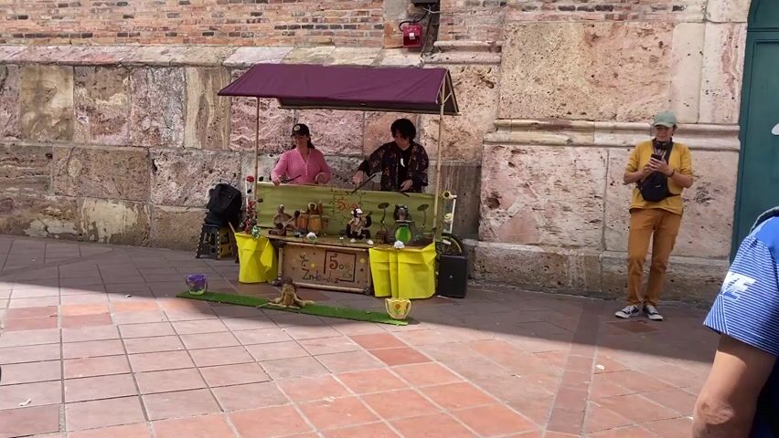 The video shows two women operating a puppet show booth on a brick-tiled plaza. The booth is covered with a maroon awning, and the counter is decorated with yellow fabric and a sign that reads "Zin-ka-dez, 5". Several small puppets are arranged on the counter, along with miniature musical instruments. One woman, wearing a pink shirt and a baseball cap, is operating the puppets while the other, wearing a patterned jacket, is playing a small drum set. Several people are watching the performance. One person is holding a small child, another is looking at their phone, and another is carrying a backpack and several objects. The booth is set up against a stone wall. The area is brightly lit by sunlight.