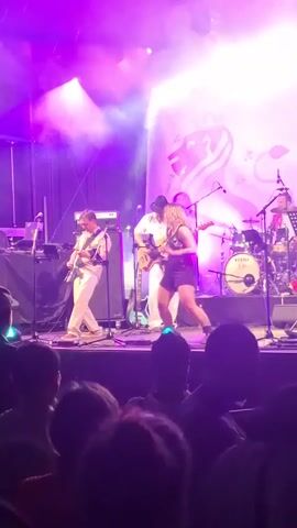 A live band performs on stage under bright purple and white lights. A female singer in a dark outfit is at the front of the stage, singing into a microphone. To her left, a male guitarist in a light-colored shirt and pants plays a guitar. Behind the singer and guitarist, another male in a white shirt and pants plays a guitar. To the right of the singer, a drummer plays a drum set, and another person appears to be playing a keyboard or synthesizer. The stage is set up with amplifiers and musical equipment. In front of the stage, the silhouettes of an audience can be seen. A large banner with a decorative design, possibly a crest or logo, is visible behind the band. The overall atmosphere appears to be energetic and festive.