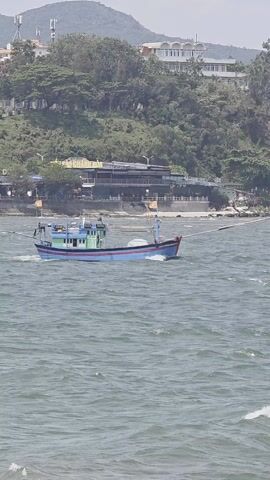 A medium shot depicts a blue and green boat sailing left to right on a wavy body of water. The boat has two long, thin poles sticking out from its sides. In the background, a verdant hillside slopes down to a sandy beach. Structures, including a multi-story building and smaller houses, are visible among the trees. The sky is overcast, casting a soft light over the scene.