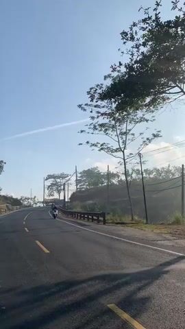 The video shows a view from a vehicle traveling on a paved road with a yellow line down the center. The road curves to the left. On the right side of the road, there is a guardrail and a grassy embankment with trees and utility poles. The sky is blue with some wispy clouds. There are two motorcycles in front of the vehicle, and a purple truck passes the vehicle on the left. The overall environment appears to be rural and hilly.