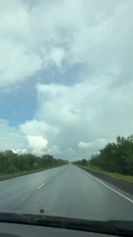 The video shows a view from inside a vehicle driving on a wet road. The road stretches into the distance, bordered by trees on either side. The sky is cloudy with patches of blue visible. A windshield wiper is briefly visible in the foreground of one frame, suggesting that it is raining or has recently rained. The overall impression is that of a drive through a rural or natural landscape under overcast conditions.