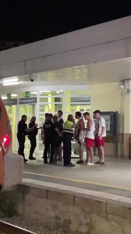 The video shows a group of people on a train platform at night. There are several uniformed individuals who appear to be law enforcement officers interacting with a group of young men. Some of the young men are wearing white t-shirts and red shorts, and appear to be in beachwear. The officers are in dark uniforms, and one officer is wearing a reflective vest. The interaction seems to be calm, with officers speaking to the young men and one officer holding what appears to be a white bag or piece of paper. In the background, there is a building with large glass doors and lighting. A train is visible on the left side of the frame, with its headlights on. The platform is well-lit, and the overall atmosphere appears to be a routine stop or interaction.