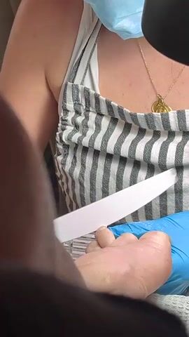 The video shows a close-up view of a person's foot, specifically the toes, being worked on by someone wearing blue nitrile gloves. A white nail file is visible, being used to shape or smooth one of the toenails. The person receiving the pedicure is wearing a striped top and a gold necklace. The scene appears to be indoors, possibly in a salon or home setting. The lighting suggests it is daytime or well-lit artificial light. The focus is on the nail treatment process.